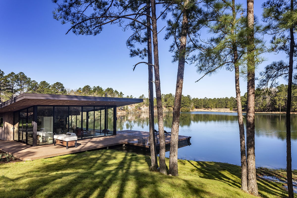 A small glass house sits on a lake, surrounded by trees.