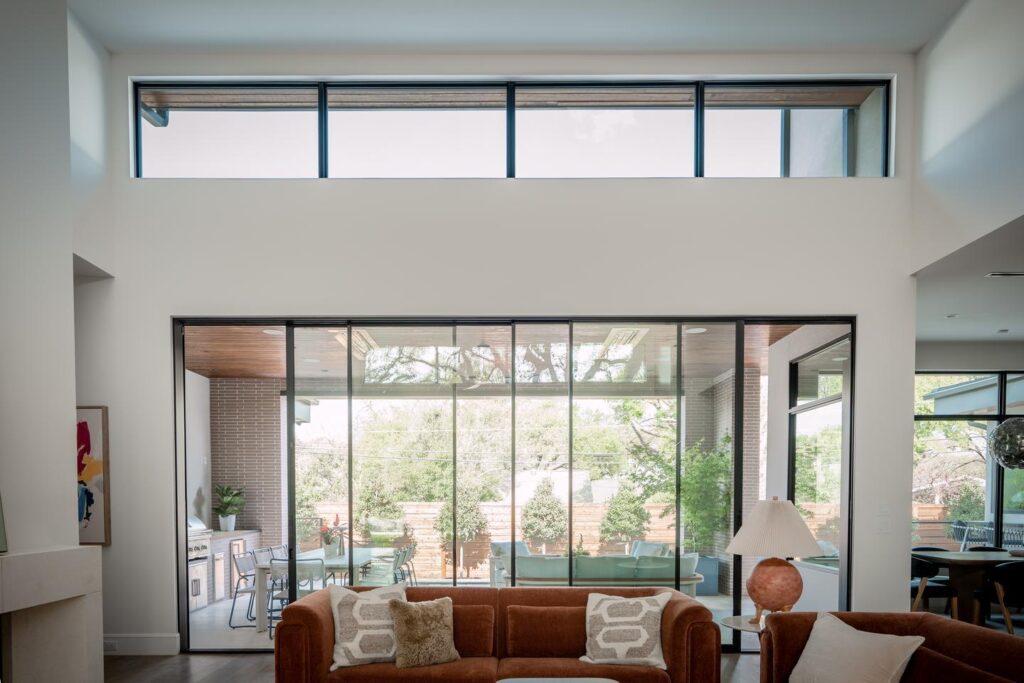 A modern interior of a residential living room shows a sofa in the foreground with a large multi-sliding door with clerestory windows above it. The backyard can be seen just outside. The whole room seems to glow from the light coming from the outside.