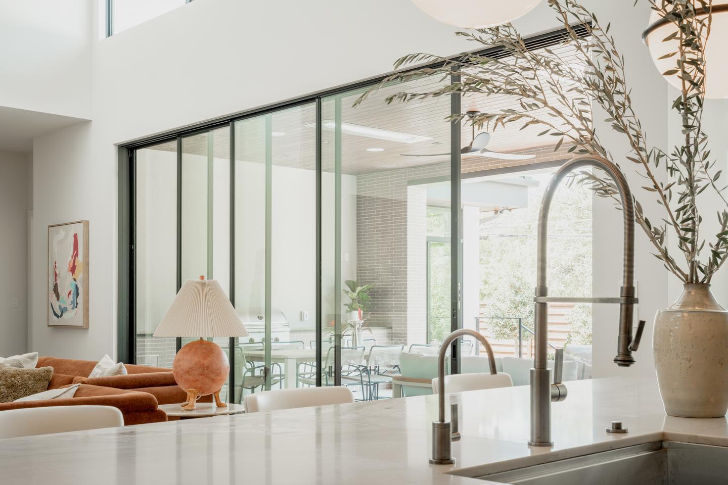 A sink and island countertop with a vase of dried grasses to the side are visible in the foreground overlooking a living room. A large multi-sliding glass door is visible on the opposite side of the room.