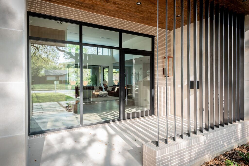A foyer and living room can be seen through a large, multi-panel window wall and glass front door. The porch includes a wood-slat wall in the foreground, which provides privacy.