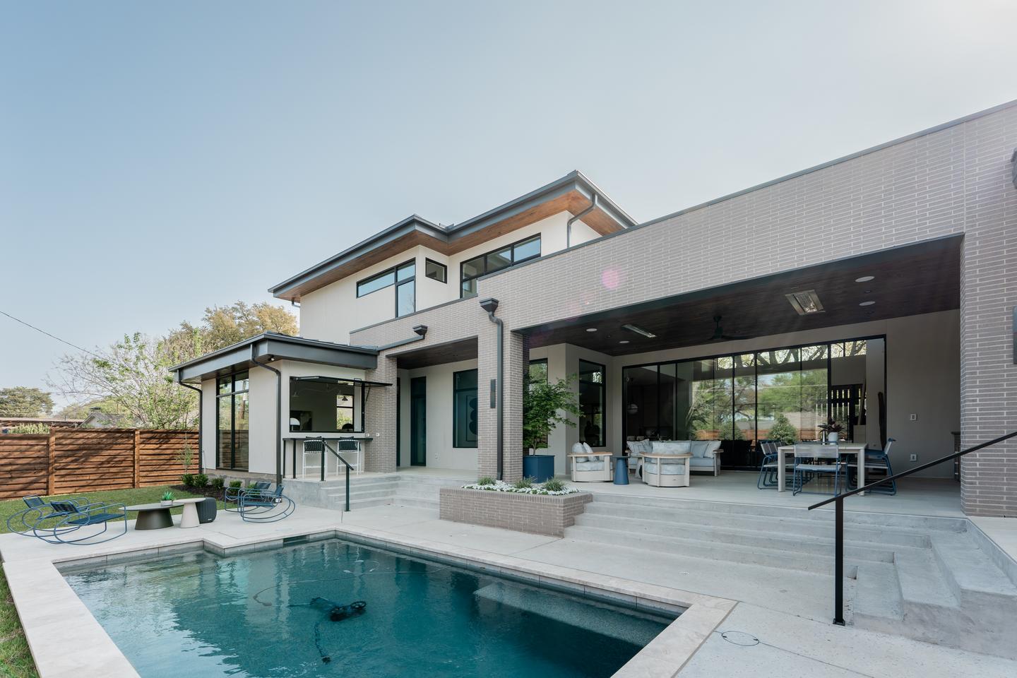 A backyard with a pool and fully furnished, covered patio of a modern home that has many large, uniquely laid out windows and doors on two levels of the home.