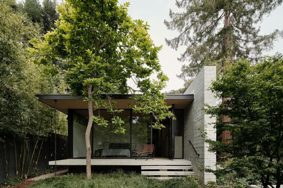 A glass wall with a Series 980 glass pivot door opens a master bedroom to a grassy, tree-filled backyard.