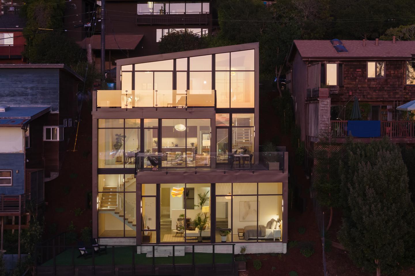 The back of a modern home on a steep hillside is entirely made of glass, and reflects the sunset while also providing a view of the layout of the home.