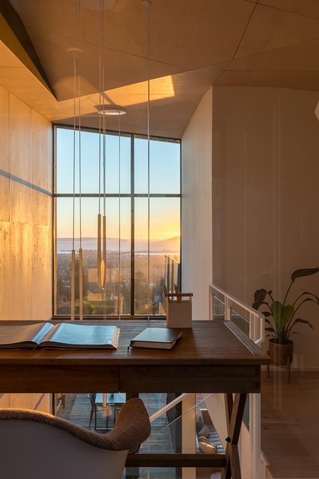 A desk overlooks an open living area and a floor-to-ceiling window wall that spans the entire opposite wall. It overlooks a bay view with the sun setting in the distance.