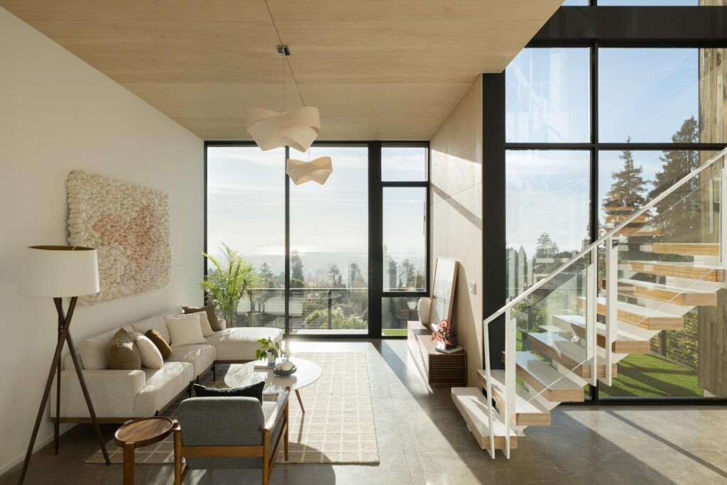A modern living room on the bottom level of a home contains a couch with a staircase opposite it. The far walls of both the room and the staircase have floor-to-ceiling glass, which allows sunlight to fill both parts of the room.