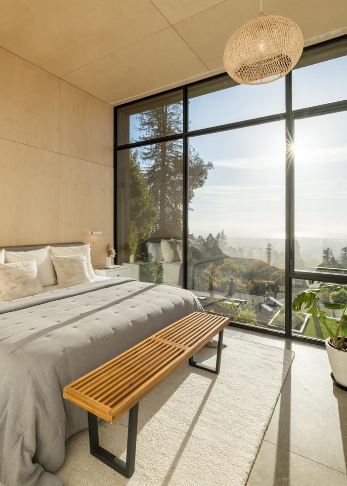 Sunlight streams through a giant window wall across a bed with a wooden bench at the foot. Trees and fog can be seen stretching into the distance.