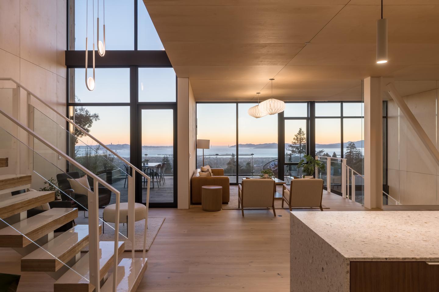 An interior of a modern home with a stone counter and stairway in the foreground, and a sitting space and living room on the far side, with a completely glass wall overlooks trees and a bay in the distance.