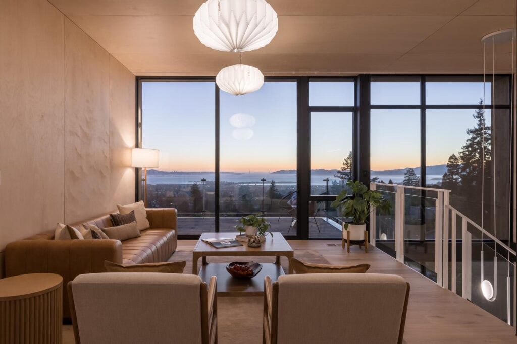 A loft in a modern home has a living room setup, with the staircase going down on the right. The sunset can be seen across a bay through the floor-to-ceiling windows that make up the entire far wall.