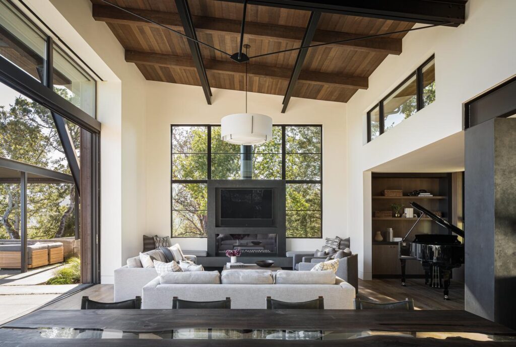 A living room with high wooden ceilings is filled with natural light from the open sliding glass door that makes up most of the wall to the left, a window wall surrounding a large TV directly opposite the viewer, and clerestory windows above the connecting room to the right.