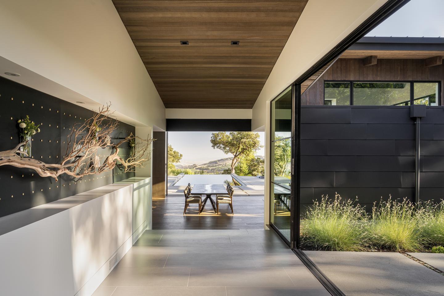 A hallway with a large open sliding glass door on the right, and a decorative branch along the left wall, leads to a dining room, and another open sliding glass door at the far end, which opens to a pool and back patio overlooking a natural scape of trees and hills.