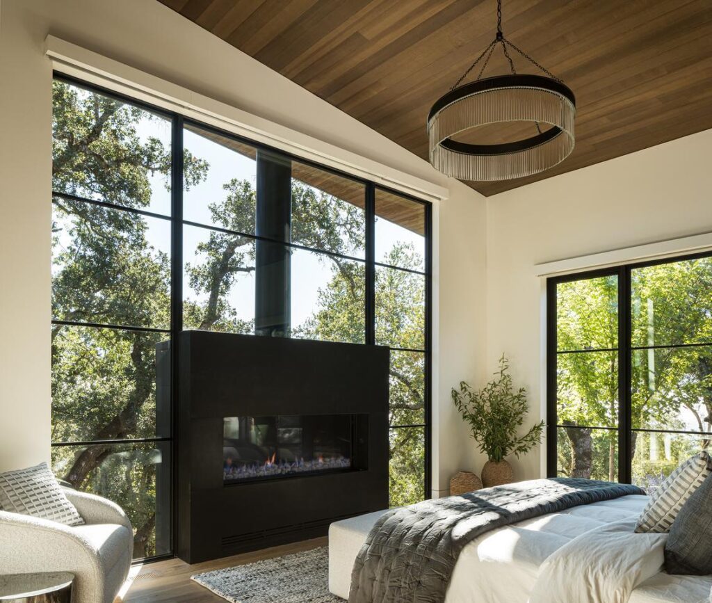 A bedroom is flooded with natural light from the giant window wall that frames a fireplace opposite the bed. Trees and sky are visible through the glass of the window wall as well as the sliding door on the adjacent wall.