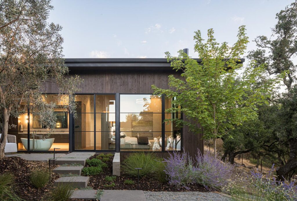 A bedroom and bathtub can be seen through a series of connected fixed windows and a sliding glass door that make up the entire length of the wall. There are steps leading up to it, and decorative trees and shrubs framing it.