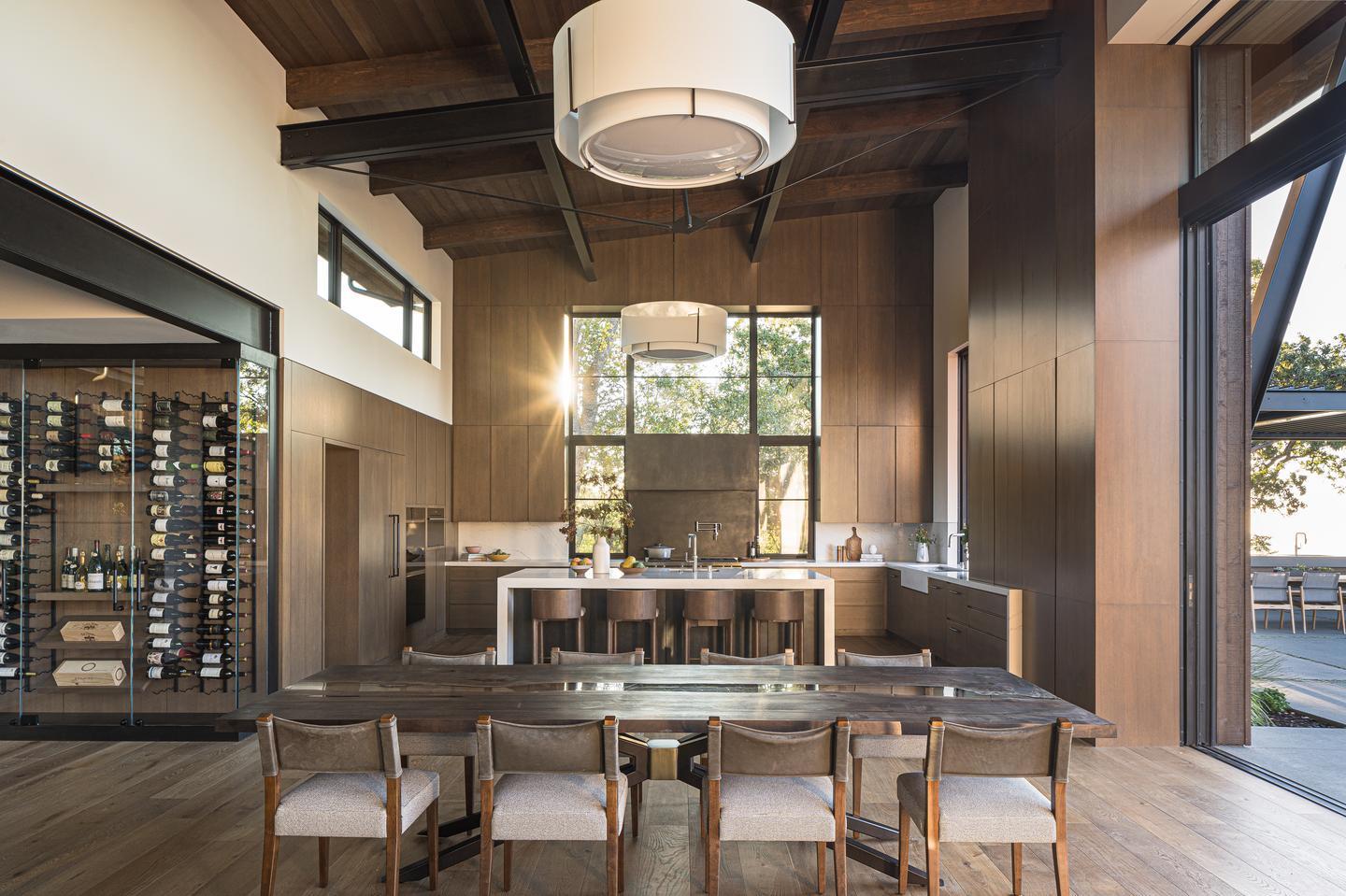 A dining room table is in the foreground with a kitchen behind it. The warm wood paneling is illuminated by the windows on the far wall surrounding the stove, as well as on the far left and right walls of the kitchen. A large open sliding door is just visible to the right of the dining table, and extends toward the viewer, out of sight.