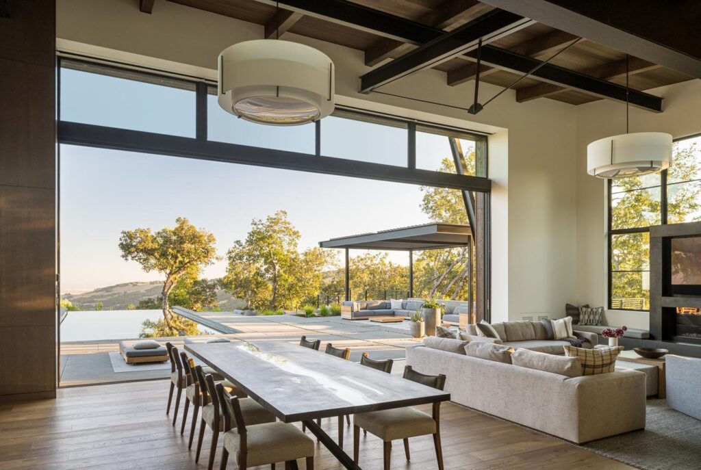 A covered outdoor pavilion and trees are visible through a large sliding door. A dining room table and sofa are in the foreground inside of the home.