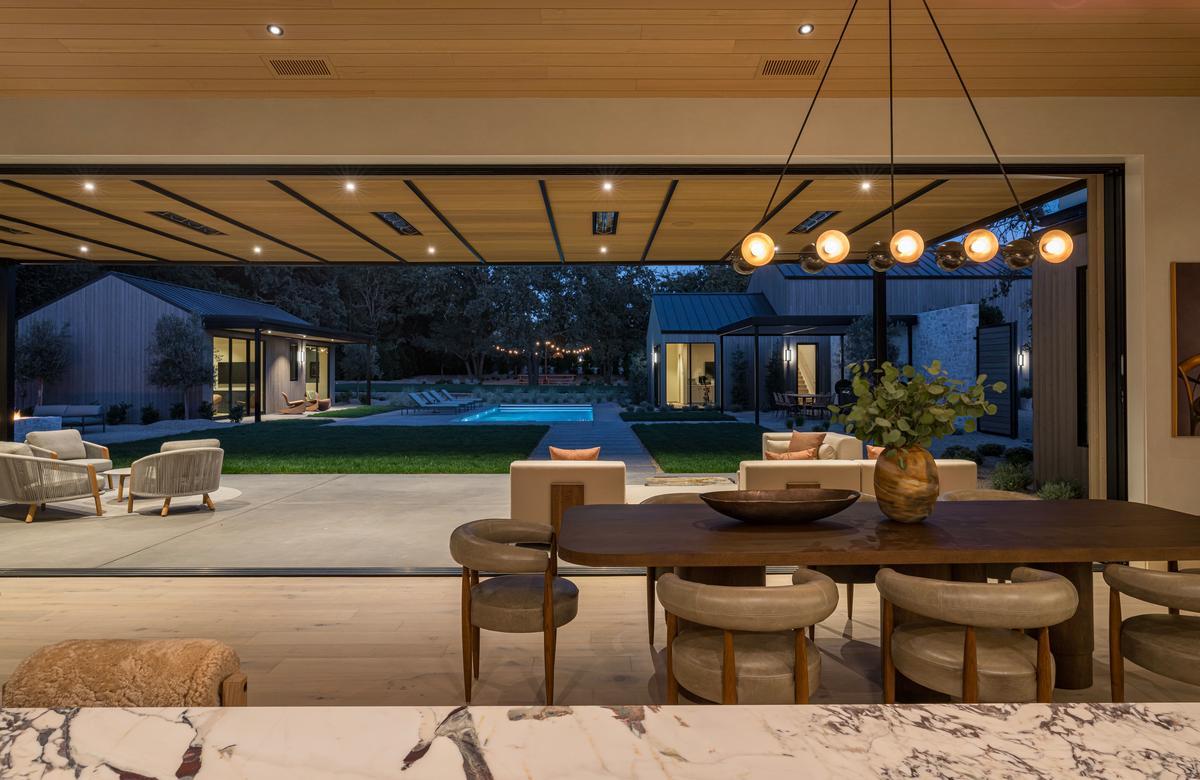 A patio and courtyard at dusk can be seen through a large opening for a sliding door with a dining room in the foreground.