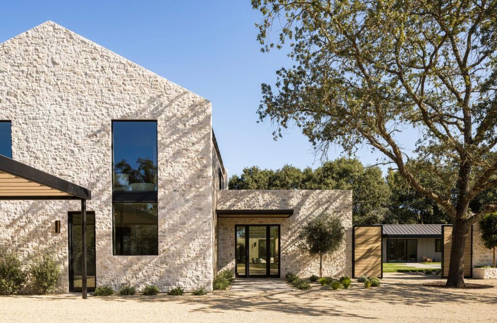 A white stone façade displays a tall window that stretches from the ground floor up to the second story. A glass entryway a little further back is shaded by a tree in the yard next to it.