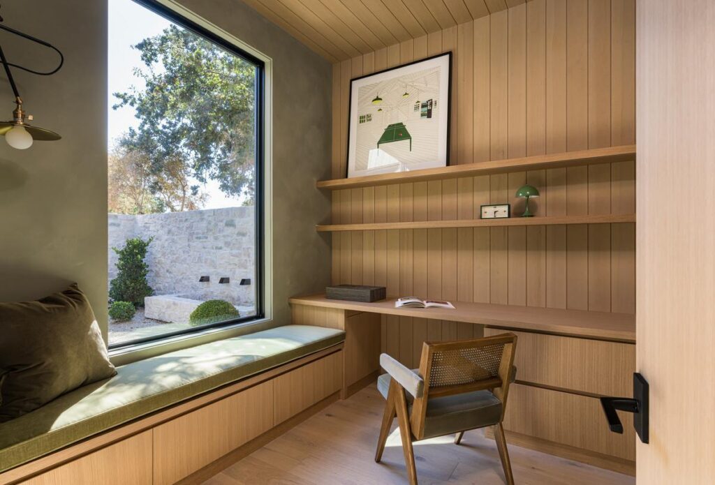 An office full of warm wood paneling and surfaces has a large fixed window with a bench seat beneath along the full length of the wall. There is a chair angled slightly towards the window.