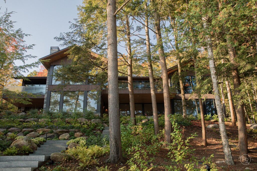 A large home in the woods with large glass windows that reflect the surrounding trees is visible behind a row of fir trees.