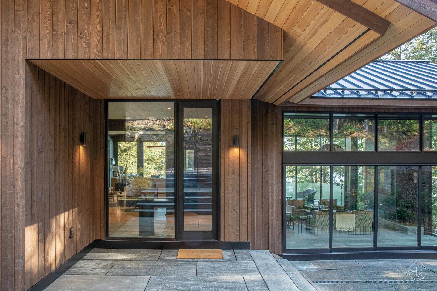 A wood-paneled entryway houses a large glass front door next to a single panel floor-to-ceiling glass window. The door, window, and a multi-slide door to the right, show the modern yet cozy interior of the home, and the trees and lake on the other side.
