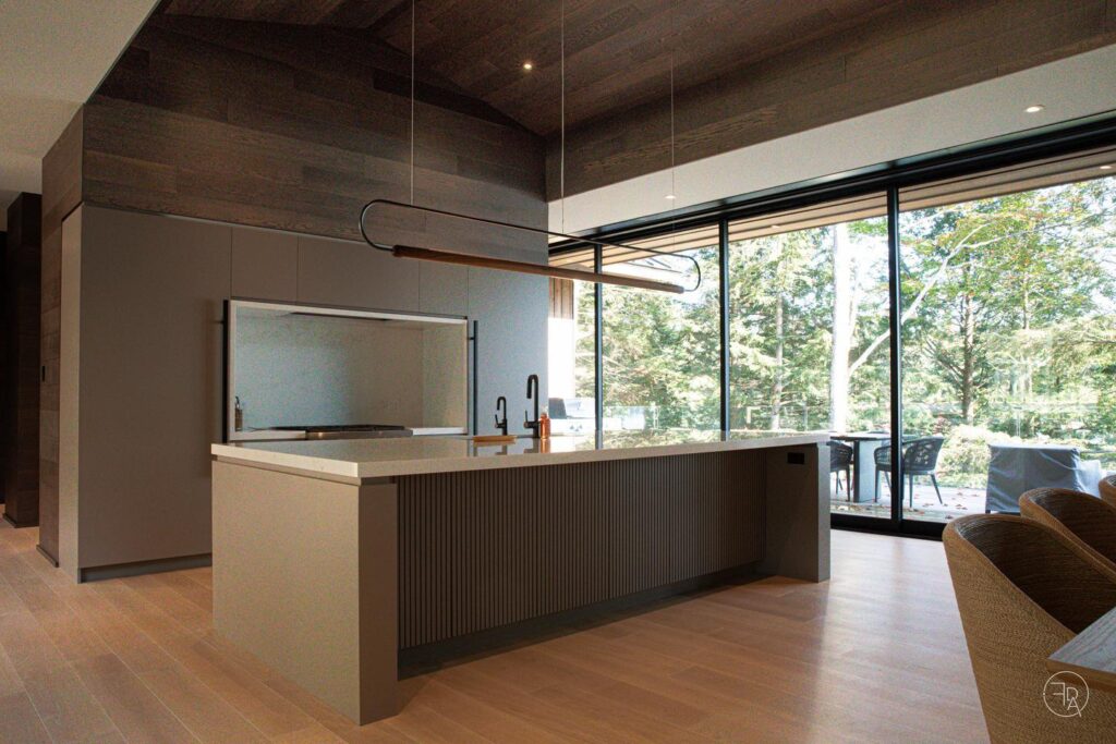 A modern kitchen designed with light natural tones, has a large floor-to-ceiling multi-slide door that overlooks a deck and a forested backyard.