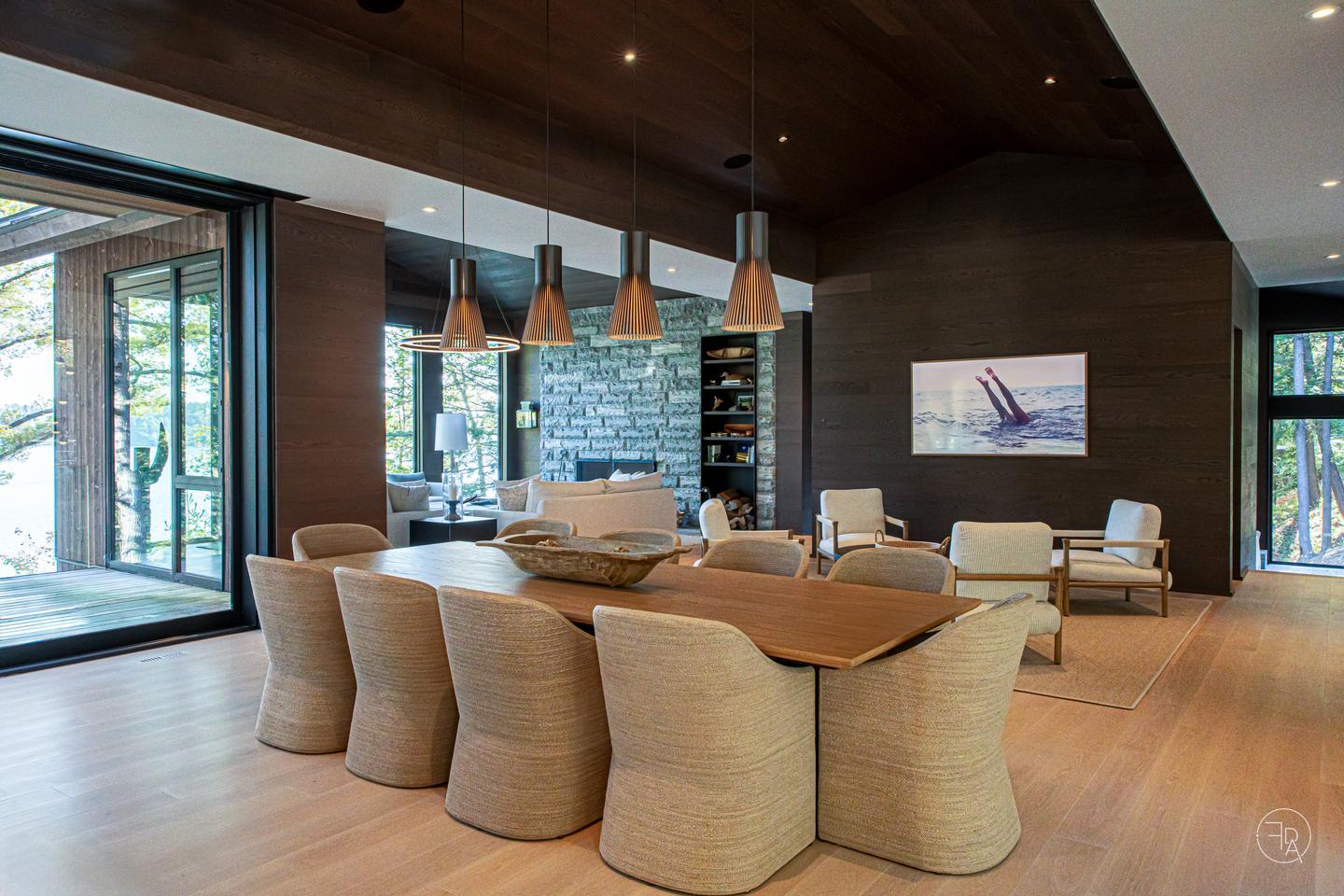 A large dining table in the foreground sits in an open-concept space. There is a sitting room with four armchairs facing a coffee table just beyond it and another wing of the home beyond that. Every wall is full of large, floor-to-ceiling windows, flooding the spaces with cool, natural light.