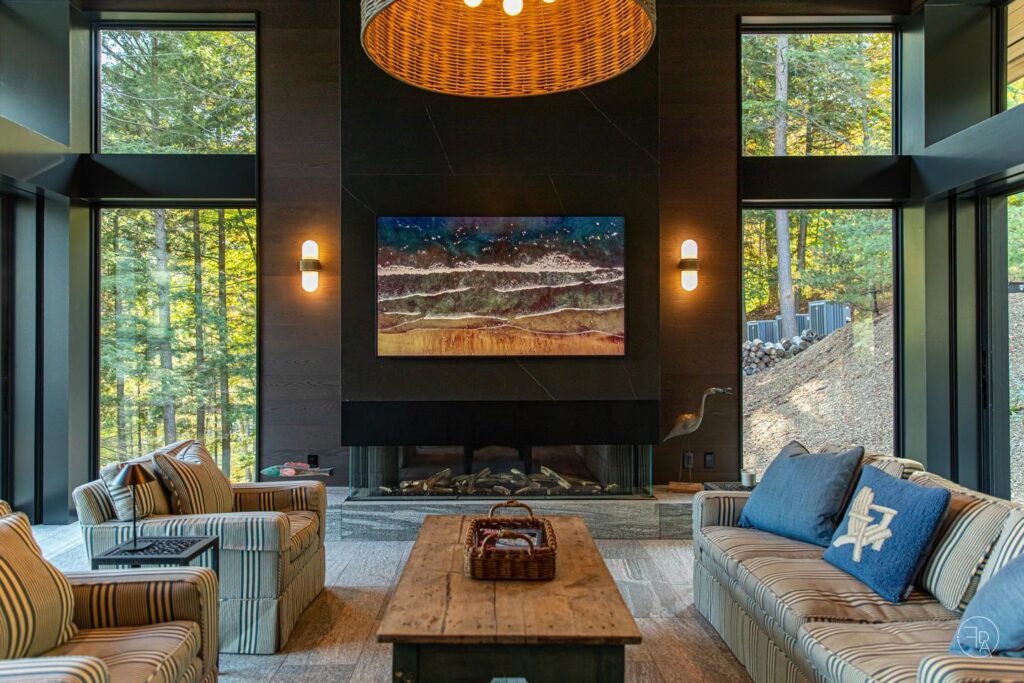 A living room in a home in the woods has two tall windows on either side of a fireplace with a large TV. The windows show the fir trees outside, and inside, a sofa sits opposite two armchairs, with a coffee table between them.
