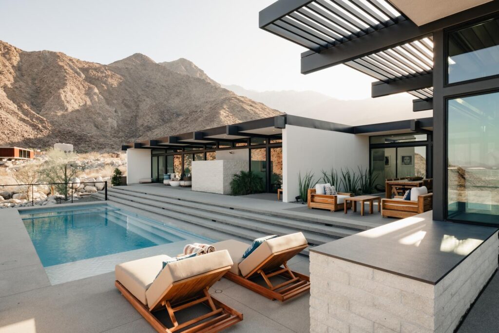 The back of a modern desert home has multiple floor-to-ceiling sliding glass doors that open onto a patio with steps that lead down to a pool. Mountains and foothills create the dramatic setting ever-changing backdrop.