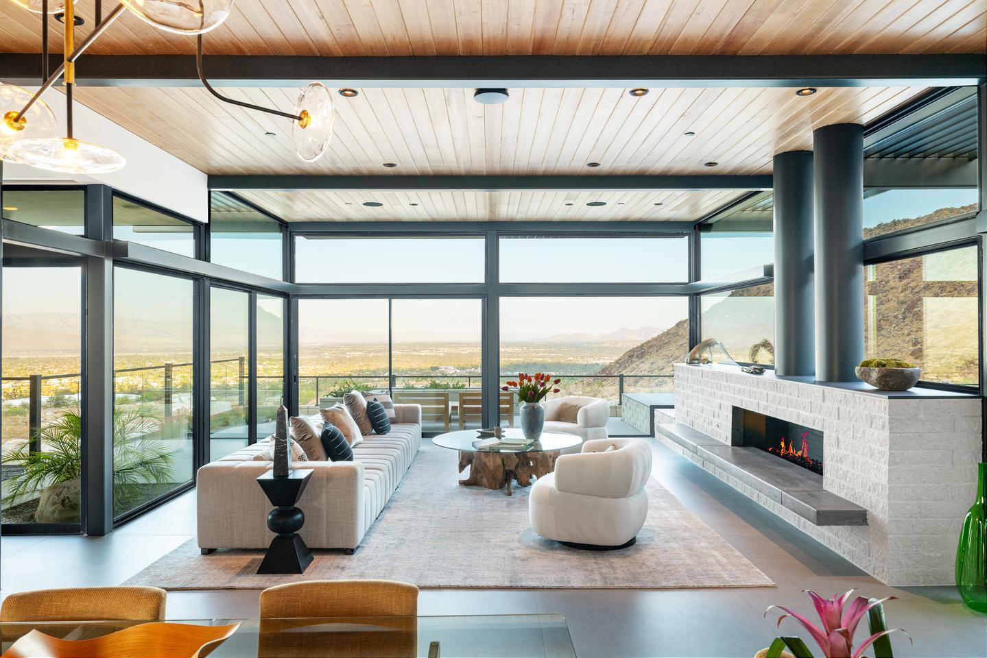 A modern living room with a fireplace and floor-to-ceiling windows and sliding glass doors overlooks a desert valley during sunset.