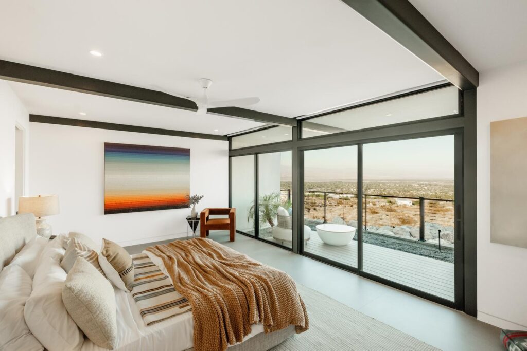 A bedroom leads to a patio through a floor-to-transom sliding glass door and transom to ceiling clearstory above all framing the desert valley beyond.