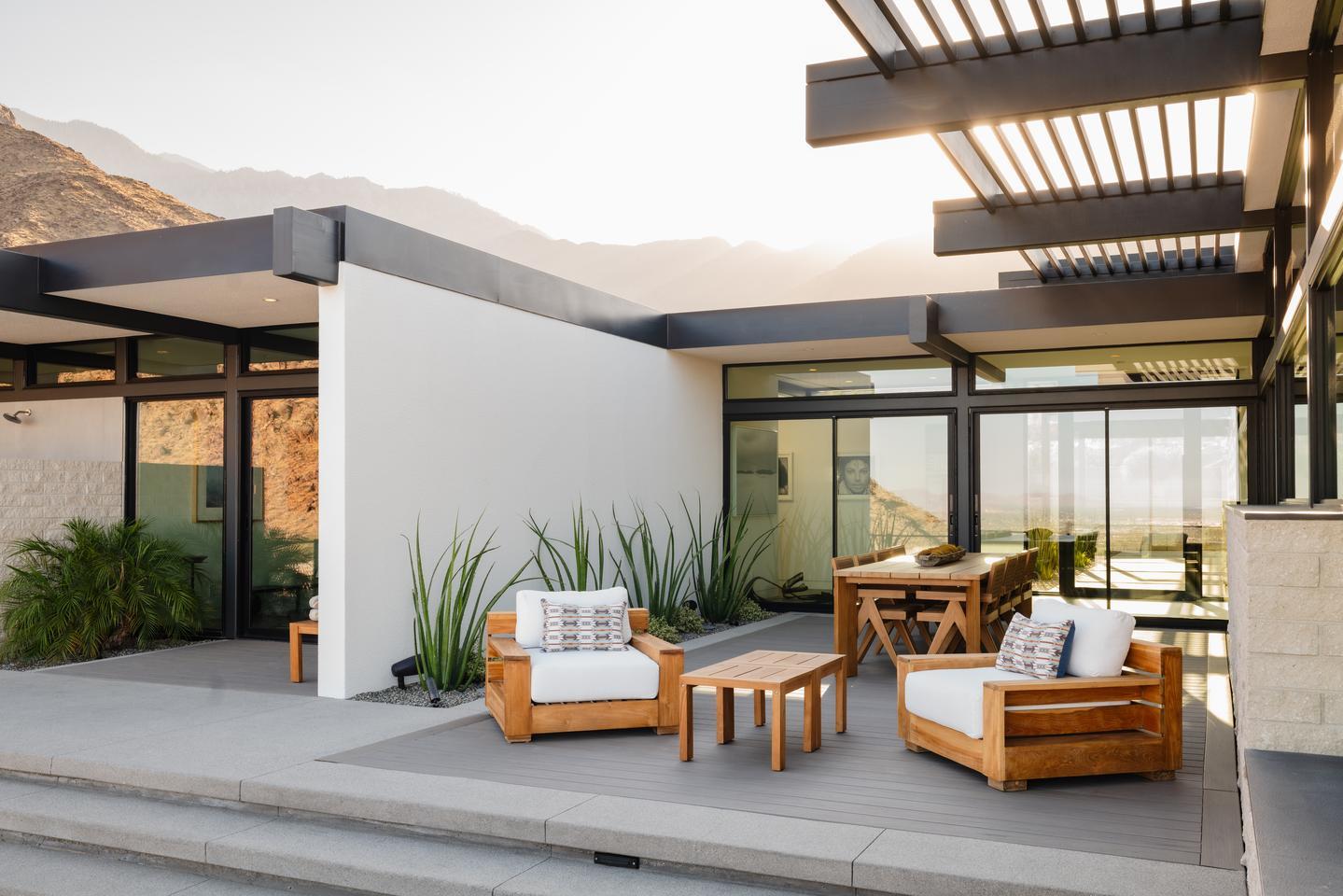 A planted patio furnished with modern, comfortable furniture, is surrounded by glass and stucco walls, which are strategically placed to filter the desert sun that is setting behind the mountains in the background.