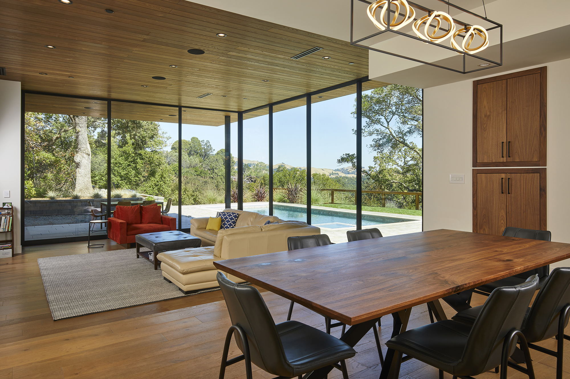 A dining room table is in the foreground of an open concept modern home. Living room furniture in front of a 90-degree multi-slide glass door can be seen in the background, with a pool and trees beyond it.