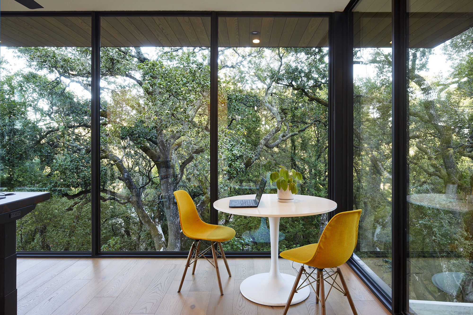 An intimate table and two chairs are in the corner of a room that is completely encased in floor to ceiling glass walls. The room is surrounded by trees.