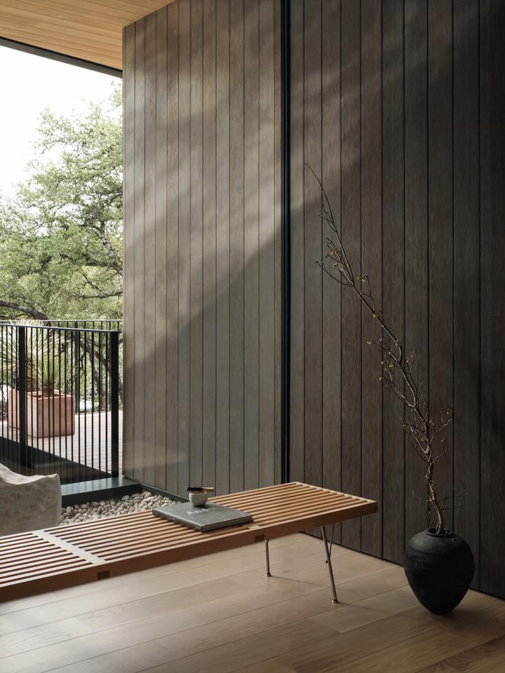 A bench is positioned in a corner in front of a fixed window that extends from floor to ceiling and from wall to wall, and looks out from the home to a rock garden and balcony.