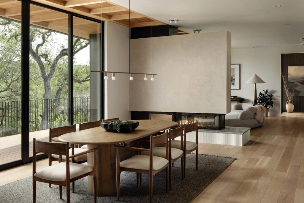 A modern dining room with natural accents leads to a balcony through a floor-to-ceiling multi-slide door. The balcony overlooks trees and a lake is visible through them.
