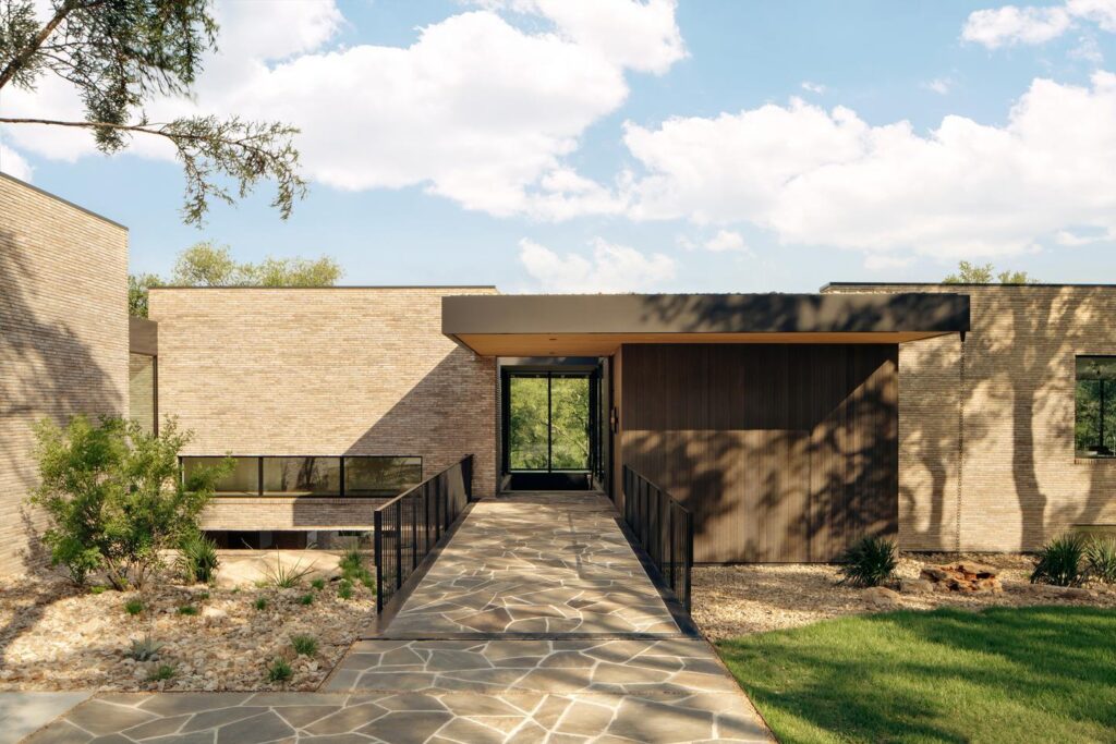 A walkway extends to a glass entryway of a tan brick home, the glass on the opposite side allows the trees behind the home to be seen.