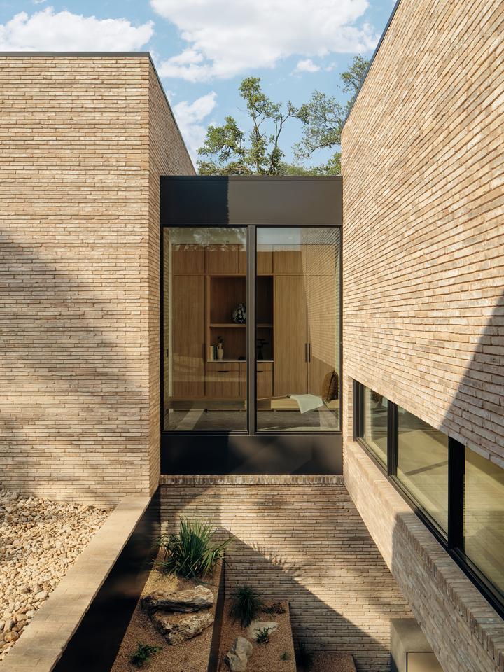 A hallway of a brick building is visible through a large glass window that is positioned above a tiered garden descending into a courtyard below ground level.