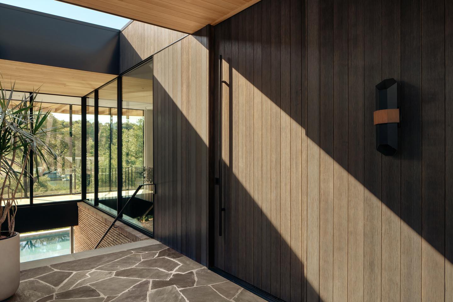 A beam of light shines diagonally across wood siding. Connected to the wood panels are glass panels, which enclose a room, and trees are just beyond the exterior glass wall.