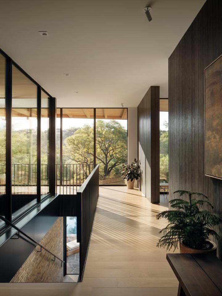 A hallway surrounded by glass walls looks out to trees. A wooden wall with a doorway is to the right, and a staircase leading down to another glass wall is on the left.