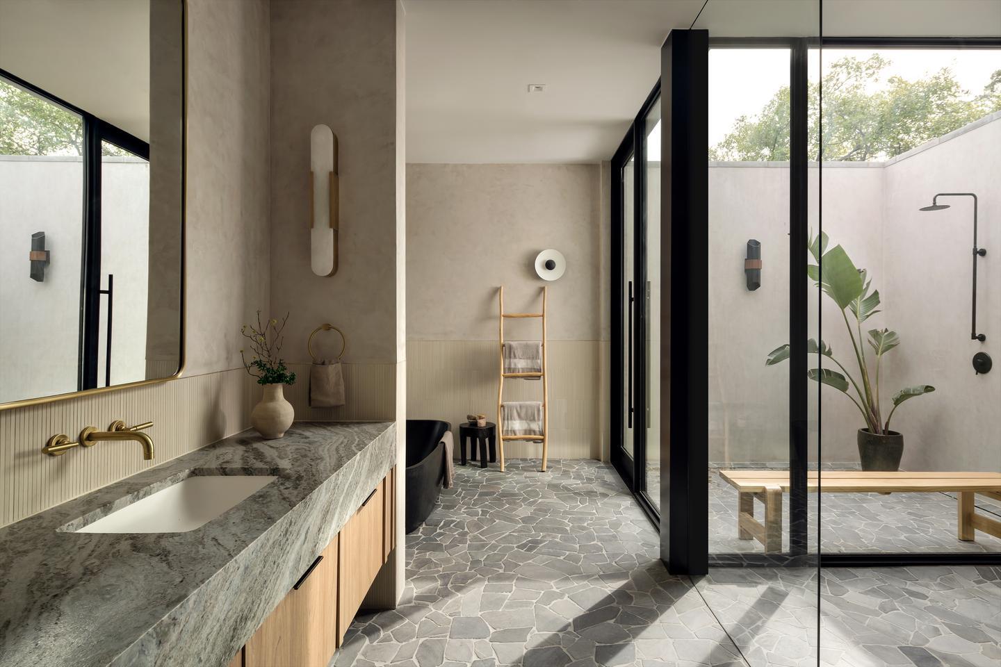 A bathroom with natural elements, sink in the foreground on the left, and a bathtub just barely visible on the other side of a wall next to the sink. To the right, an outdoor shower is visible through the floor-to-ceiling glass walls.