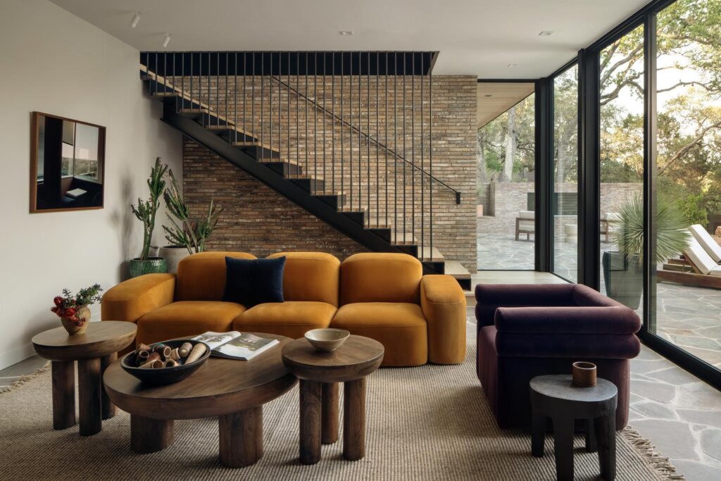 A sitting room with modern furniture is surrounded by walls built with natural materials and colors, and includes a sliding glass door that leads to an outdoor area.